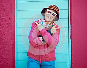 Portrait, peace and funky with a senior woman outdoor standing against a blue door and red wall background. Glasses
