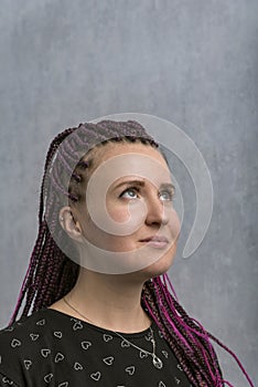 Portrait of pencive young caucasian woman looking up, with violet artificial dreadlocks on gray background