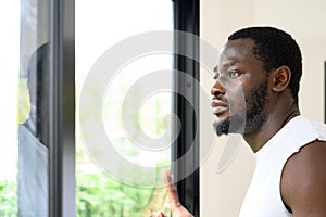 Portrait of pensive African American man standing, touching, and looking outside the window at home