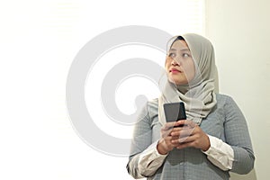 Portrait of a pensive asian muslim businesswoman using mobile phone and looking away, standing near window on white background