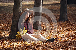 Portrait of pensive brunette girl in autumn fall park in brown hat, sweater and trousers. woman sitting on leaves near