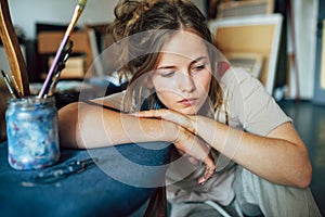 Portrait of a pensive female artist sitting on the floor next to the chair in her art studio. A woman painter posing in the