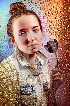 Portrait of pensive girl with black tulip behind window glass with drops of rain and colorful reflection from rainbow or