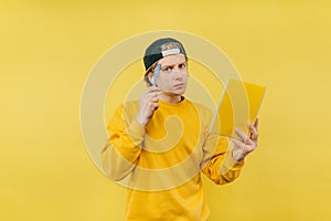 Portrait of a pensive guy in a cap with a notebook in his hand and a pen standing on a yellow background and looking at the camera