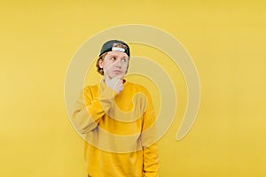 Portrait of a pensive guy in a sweatshirt and cap stands on a yellow background and looks away and thinks