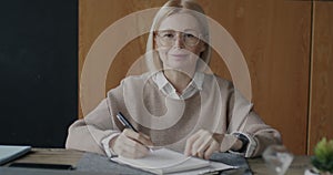 Portrait of pensive mature woman writing notes then looking at camera and smiling indoors in modern office