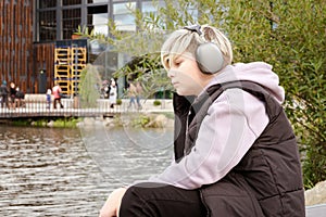 Portrait of pensive sad caucasian teenager boy sitting on the lake shore and listening to music on wireless headphones