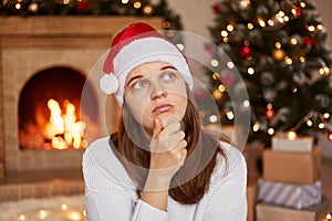 Portrait of pensive sad woman wearing white sweater and santa claus hat sitting in decorated living room, looking away, holding