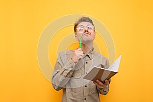 Portrait of pensive student on yellow background with exercise book and pen in hand on yellow background, looking up and thinking