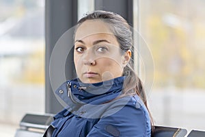 Portrait of a pensive woman 40-45 years old, sitting in a glass bus stop, waiting for a bus, looking away.