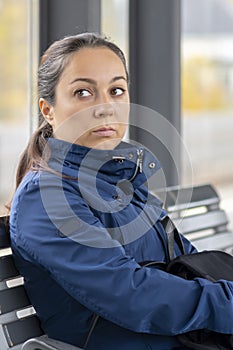 Portrait of a pensive woman 40-45 years old, sitting in a glass bus stop, waiting for a bus, looking away.