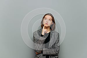 Portrait of a pensive young business woman in a jacket, standing on a gray background, looking up and thinking