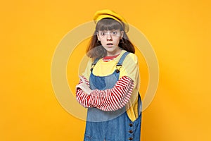 Portrait of perplexed girl teenager in french beret, denim sundress holding hands folded isolated on yellow wall