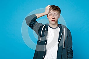 Portrait of perplexed young man in casual clothes putting hand on head isolated on blue wall background in studio