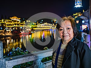Portrait photo of Asian senior woman with beautiful nightscape of fenghuang old town.