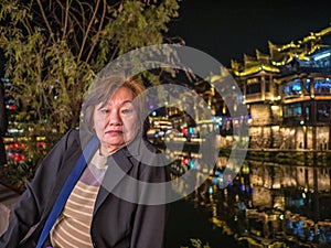 Portrait photo of Asian senior woman with beautiful nightscape of fenghuang old town.