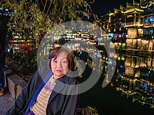 Portrait photo of Asian senior woman with beautiful nightscape of fenghuang old town.