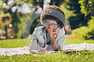 Portrait photo of cheerful smiling black skinned girl laying in the park, writing notes with pen wearing jeans outfit