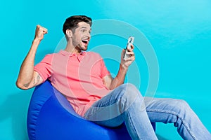 Portrait photo of overjoyed crazy young man raised fist up betting sports using smartphone sit pouf isolated on