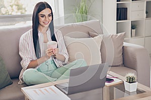 Portrait photo of young female worker using laptop at home smiling keeping cup with warm beverage wearing spectacles