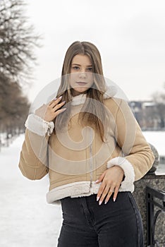 Portrait of photogenic girl in beige sheepskin coat in snow-covered park . Young woman in fashionable winter clothes