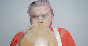 Portrait of the pink head man blowing up the orange ballon. His reaction on its popping. He is cleaning the ear with the