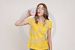 Portrait of playful brown haired teenager girl in yellow T-shirt looking at doughnut and sticking out tongue, having fun with