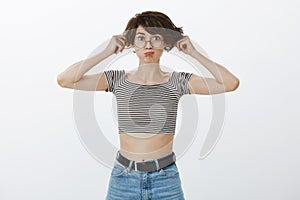 Portrait of playful childish cute woman in round eyewear and cropped striped top, pulling ears and making funny face