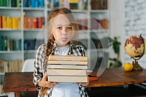 Portrait of playful elementary child school girl holding stack of books in library at school, looking at camera.