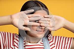 Portrait playful smiling teen girl hiding looking through hand one eye on yellow background.