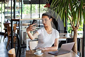 Portrait of a playful young girl taking selfie with mobile phone while sitting with laptop computer at a cafe outdoors. Morning