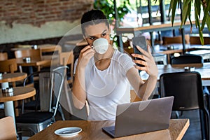 Portrait of a playful young girl taking selfie with mobile phone while sitting with laptop computer at a cafe outdoors. Morning