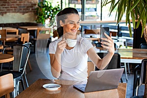 Portrait of a playful young girl taking selfie with mobile phone while sitting with laptop computer at a cafe outdoors. Morning