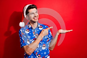 Portrait of pleasant person dressed santa hat blue shirt look directing at empy space on hand isolated on vibrant red