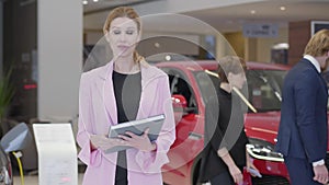 Portrait of pleasant woman in pink jacket explaining information about cars in camera holding big book close up