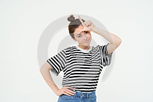 Portrait of pleased, arrogant young woman, showing loser gesture, l letter on forehead, smirking, standing over white