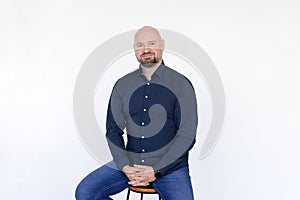 Portrait of pleased middle-aged man wear blue shirt, jeans sitting on stool, interlacing fingers on white background.