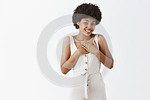 Portrait of pleased and touched cute dark-skinned female entrepreneur in trendy white overalls holding palms on chest