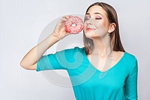 Portrait of pleasure dreaming young beautiful girl in blue blouse standing, holding and showing pink donut with closed eyes in gr