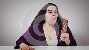 Portrait of plump Caucasian woman smelling and greedily eating sausage. Fat brunette girl hungrily biting food. Obesity