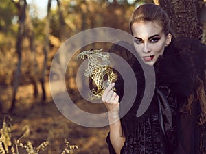 Portrait of posh dangerous lady vampire in the woods holding delicate mask and looking straight with predatory gaze.