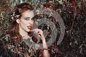 Portrait of posh woman with provocative make up and predatory gaze standing in the berry bush and eating red berries