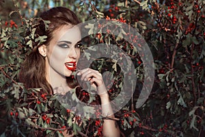 Portrait of posh woman with provocative make up and predatory gaze standing in the berry bush and eating red berries photo