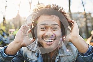 Portrait of positive adult dark-skinned man smiling broadly while sitting in park, listening to music in headphones and