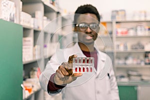 Portrait of positive African American man pharmacist in modern drugstore holding red pills in the hand. Focus on the