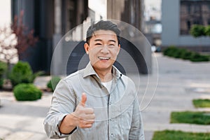 Portrait of positive asian man standing outdoors and gesturing thumb up, walking in urban city area, copy space