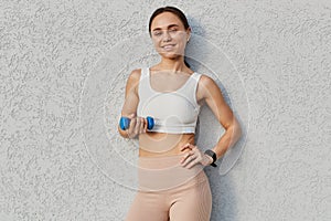 Portrait of positive attractive female wearing white sporty top and beige leggins standing with dumbbell in hand, doing sports