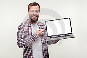 Portrait of positive bearded man in casual plaid shirt holding laptop and pointing at empty screen. white background, place for ad