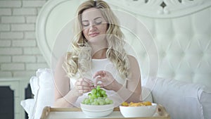 Portrait of positive beautiful pregnant woman eating healthful green grapes instead of harmful potato chips, and smiling