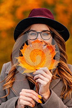 Portrait of a positive beautiful young woman in stylish glasses in an elegant burgundy hat in an autumn coat with an orange sheet